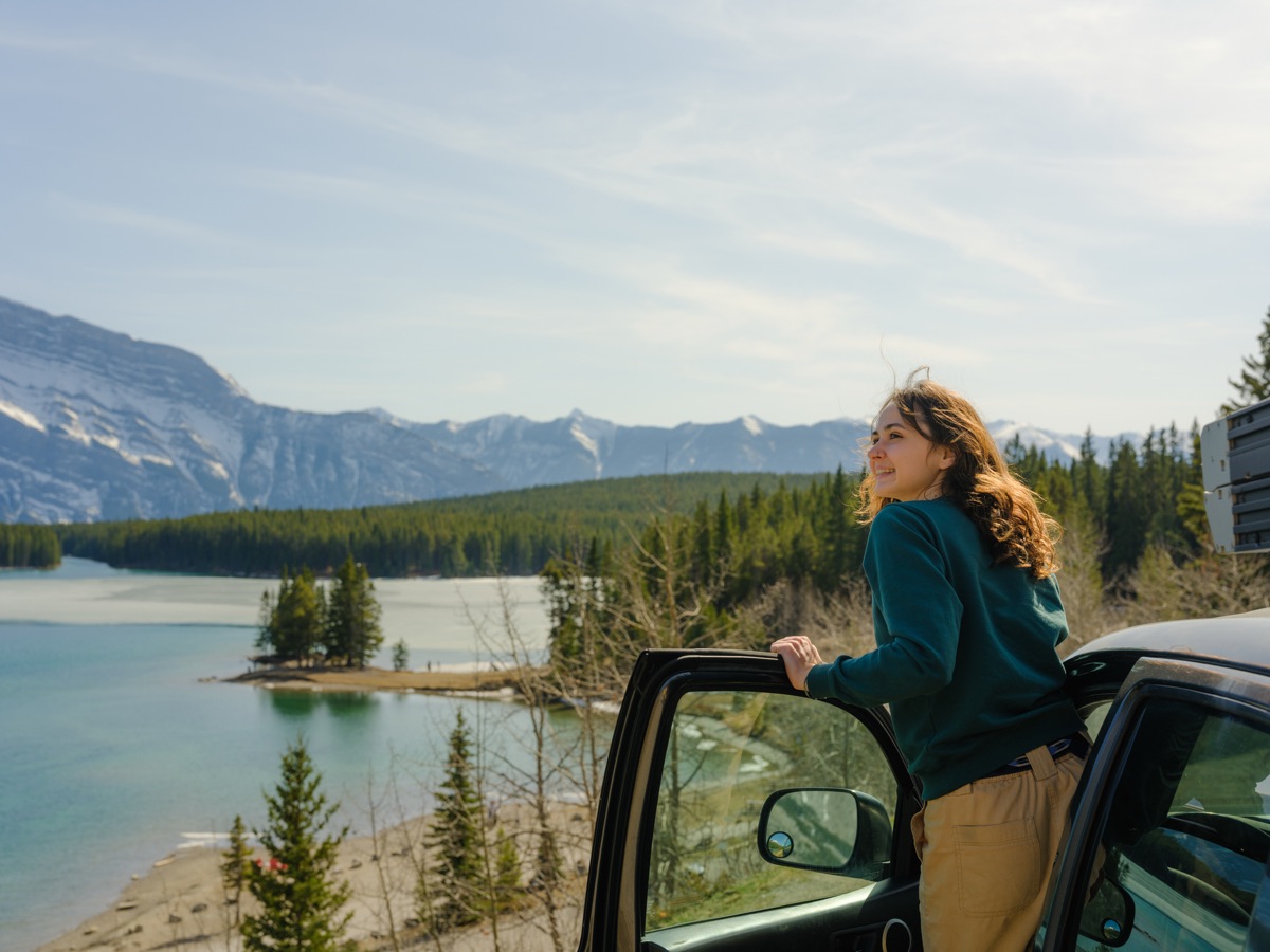 A woman leaning on an open car door, smiling at a lake below snowy mountains